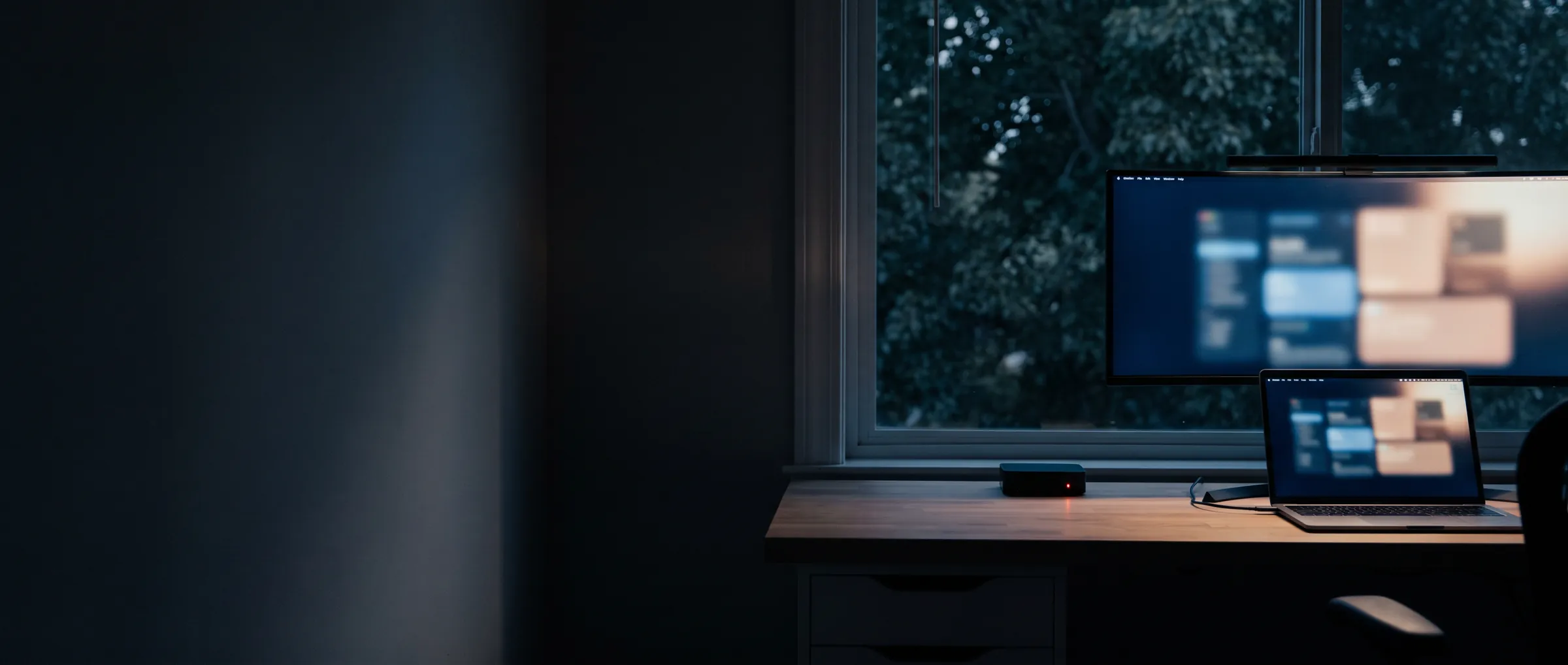 Un escritorio de noche junto a una ventana con árboles, con un monitor y una laptop que muestran un tablero de monitoreo